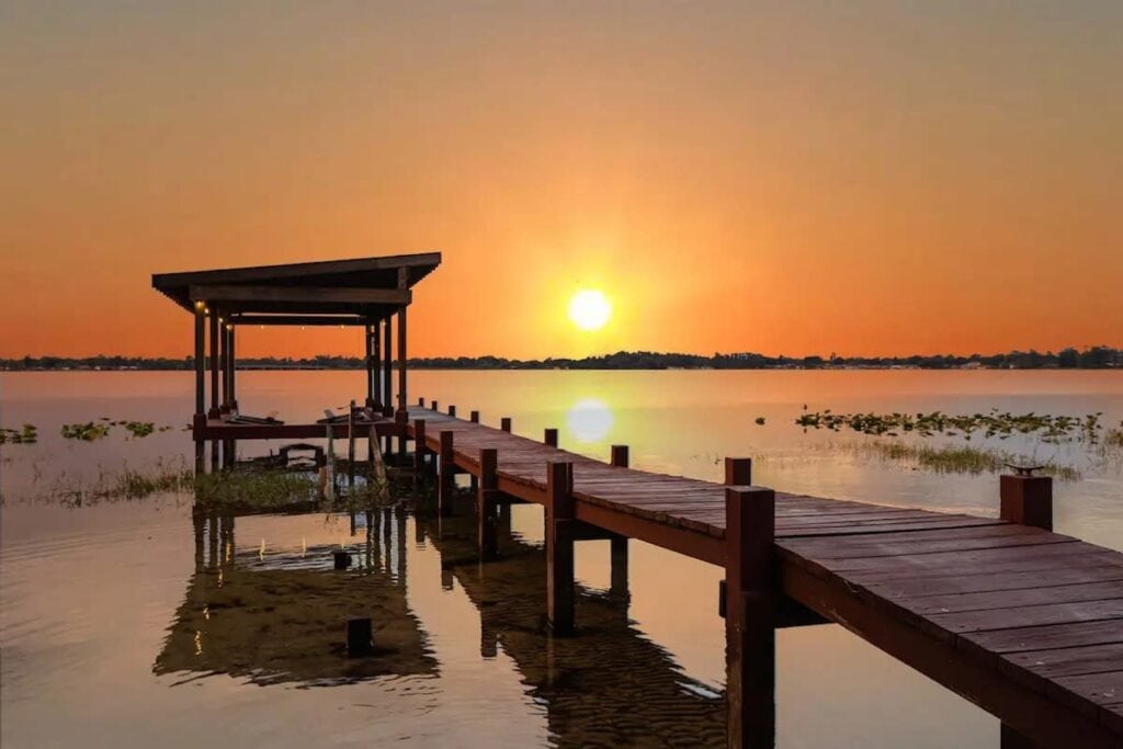 A wooden dock extends into a lake with the sun setting in the distance over the water