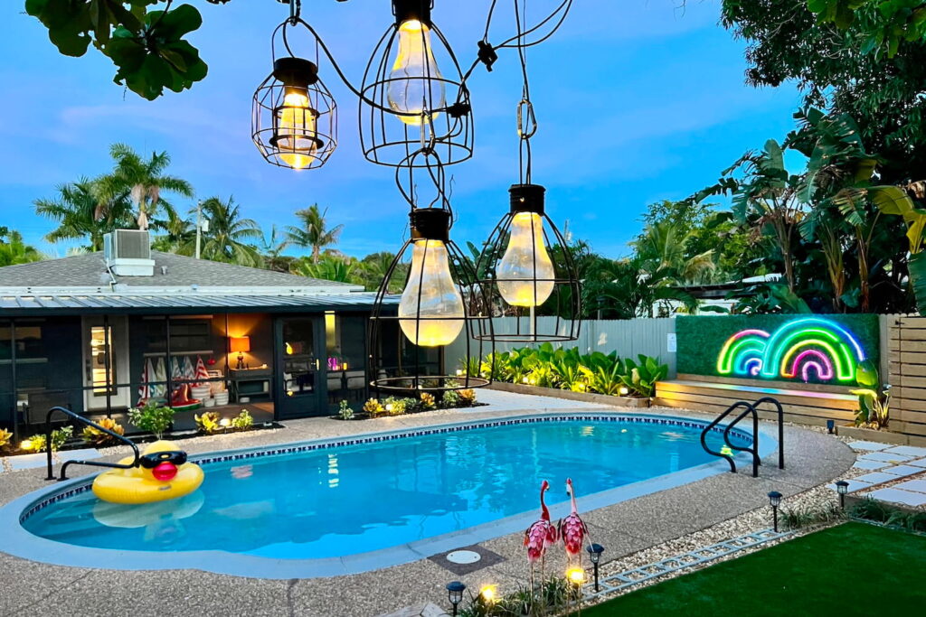 Backyard pool scene with palm trees in background, a rubber duck float in the pool, beautiful landscaping, and lots of lovely lighting, including a neon double rainbow sign