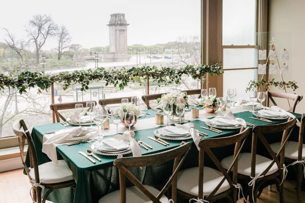 Wooden chairs surround a small reception table decorated with green and white linens and small white floral centerpieces in brass vases by a large window looking out on a park and monument