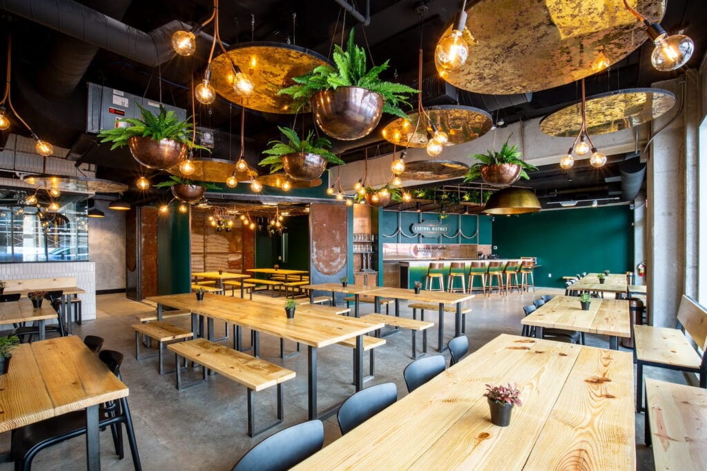 Hanging plants in bronze planters line the ceiling of a party venue decorated with long picnic tables and bar stools