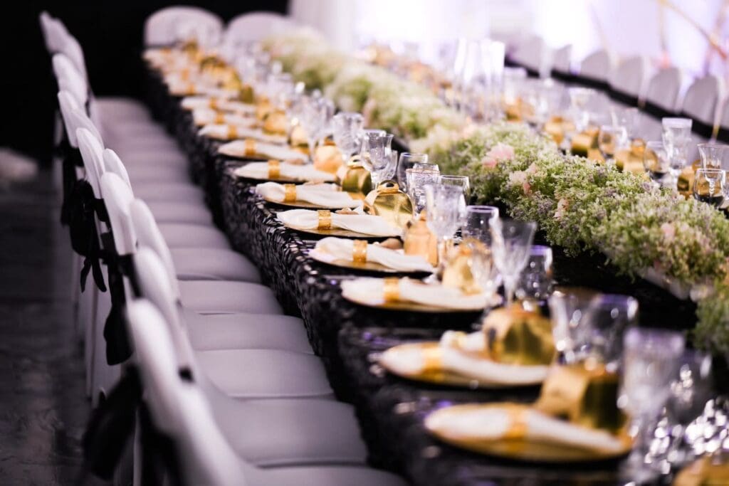 A long reception table covered in a dark tablecloth and decorated with gold accents and low-lying flowers