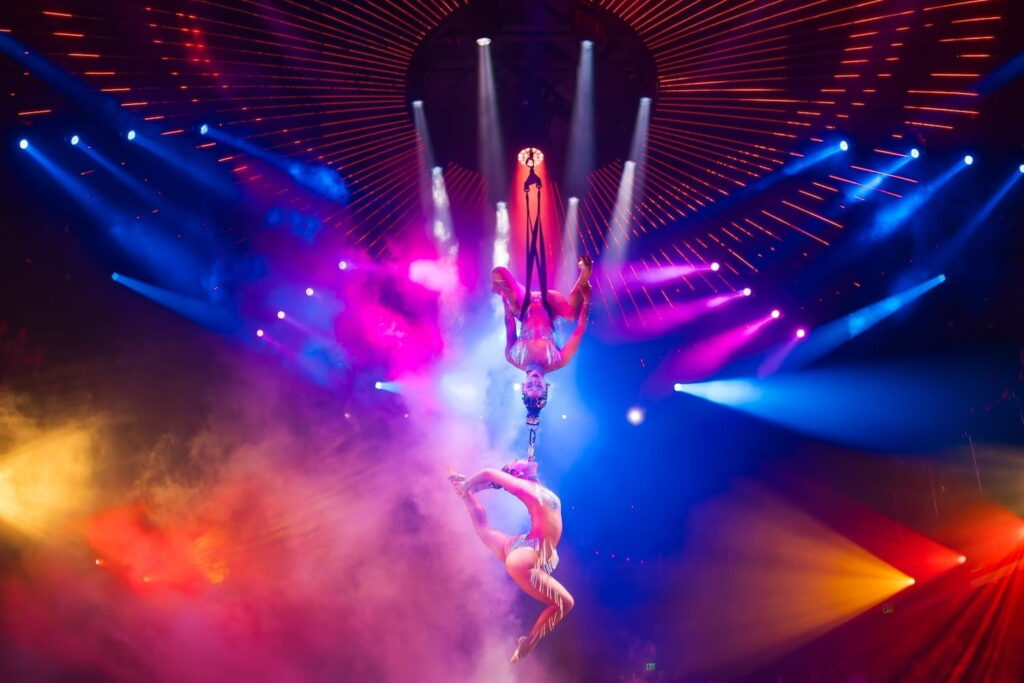 Two acrobats are suspended from the ceiling, one hanging from the other, while illuminated by neon lights