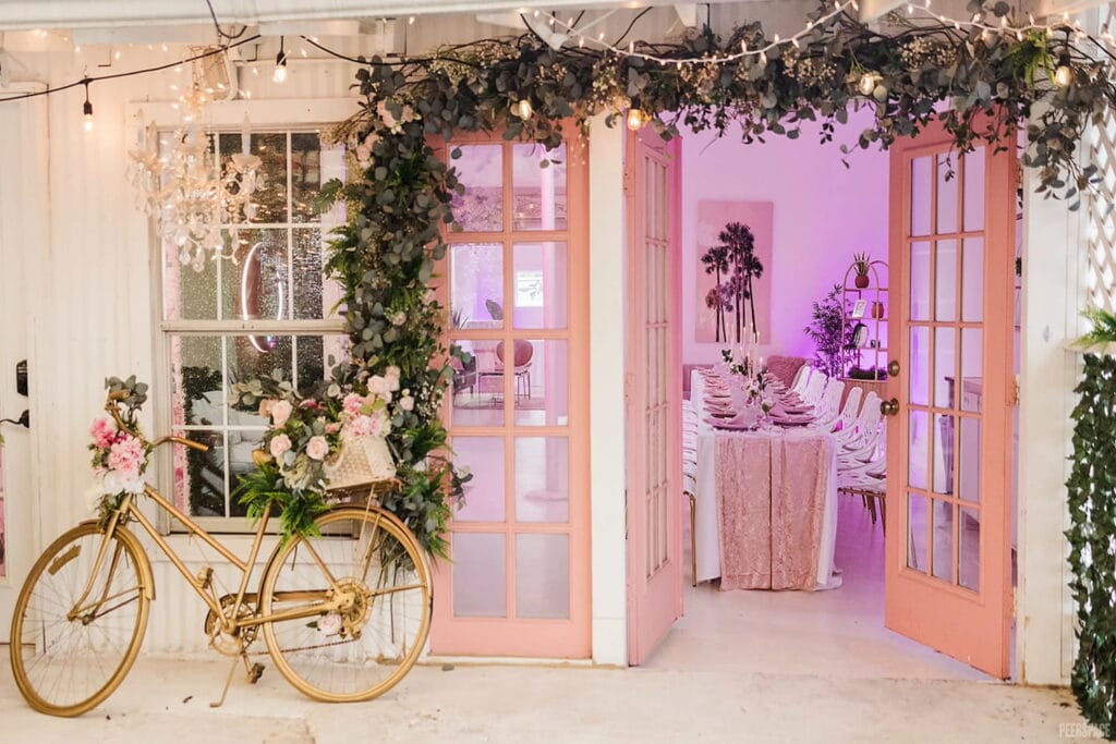 A gold bicycle decorated with flowers sits outside pink doors looking into a party space with pink accents
