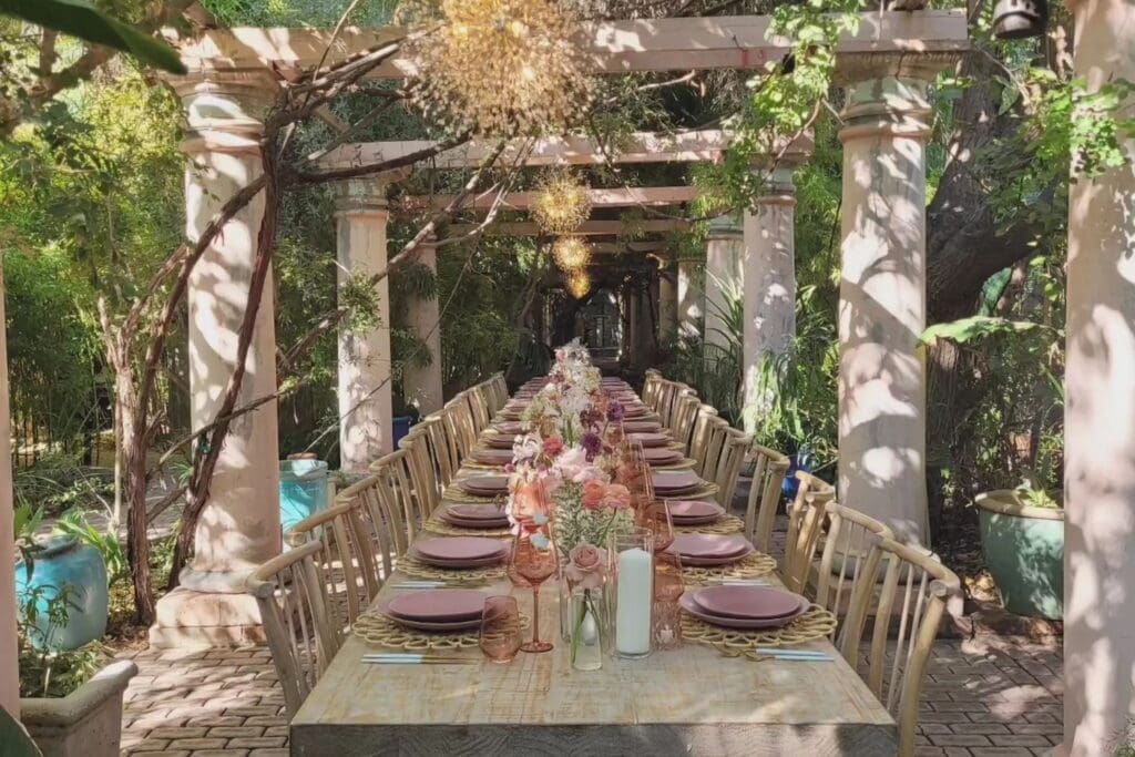 A romantic outdoor setting featuring a long wooden banquet table nestled between stone columns and decorated with mauve, peach and purple flowers and plates with sunburst chandeliers hanging above