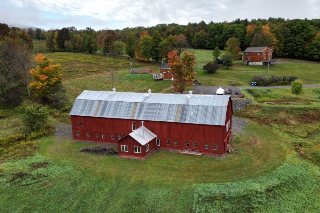 Aerial shot of a farm in New York State with a main home, small greenhouse, and large red barn set among autumn-shaded trees