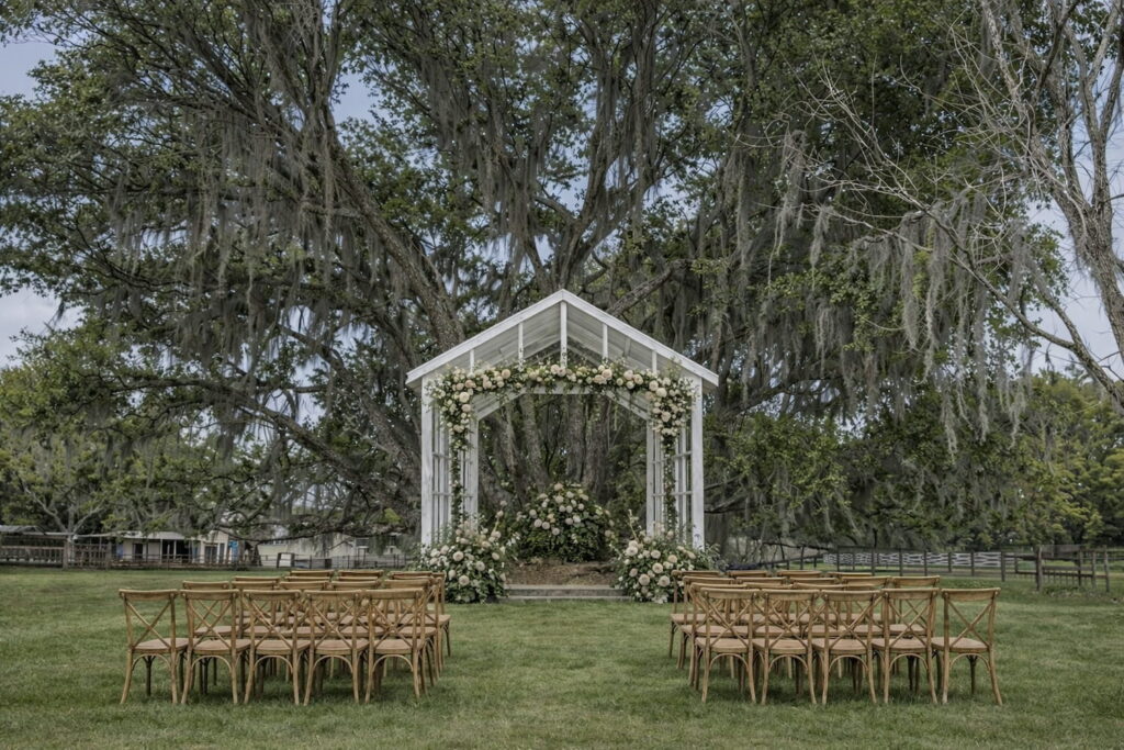 An outdoor wedding ceremony set up by a white gazebo covered in flowers with large trees in the background