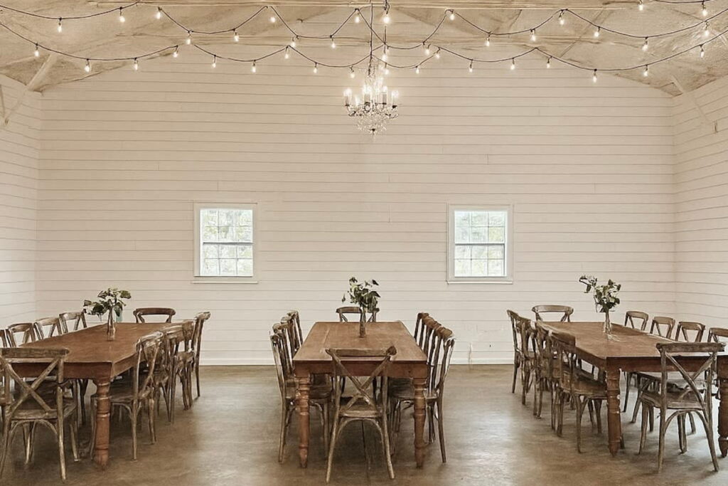 Interior of a farmhouse event space with white clapboard walls and long wooden tables illuminated by a chandelier and string lighting