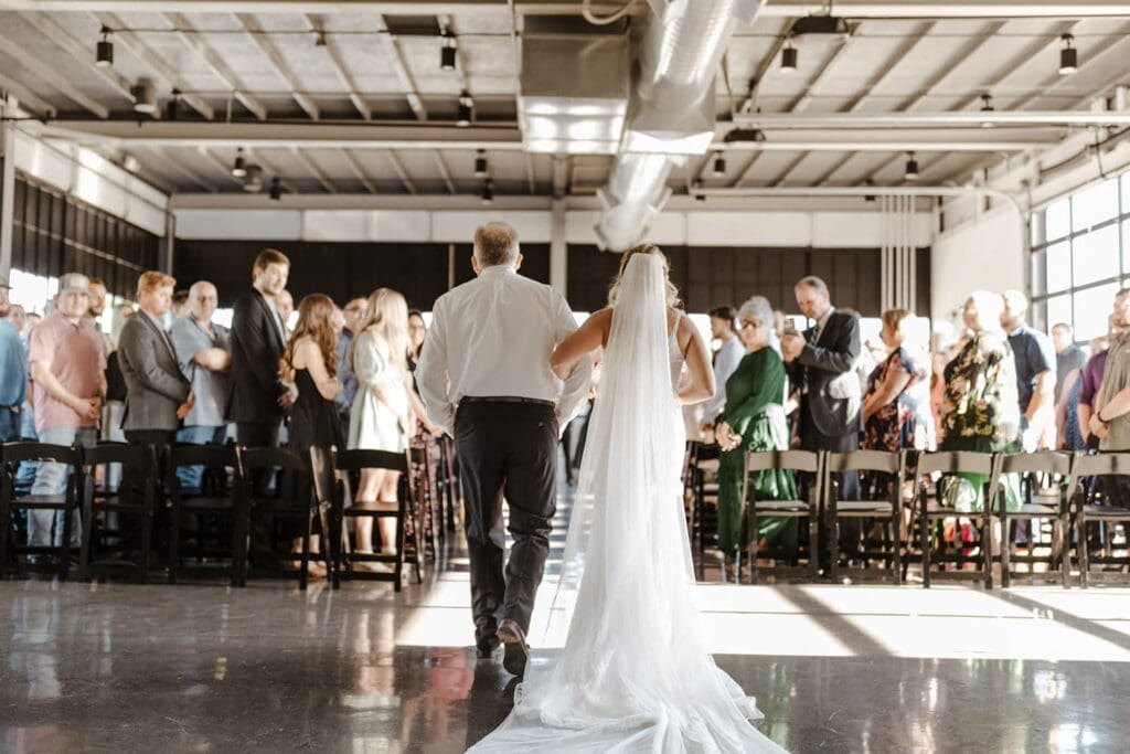 A crowd stands facing a bride and her escort as they start walking down a ceremony aisle in an industrial space