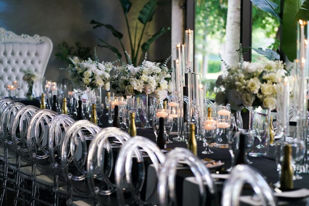 A formal reception with clear ghost chairs lining a black table decorated in black and gold bottles of champagne, white and green centerpieces and tall candles