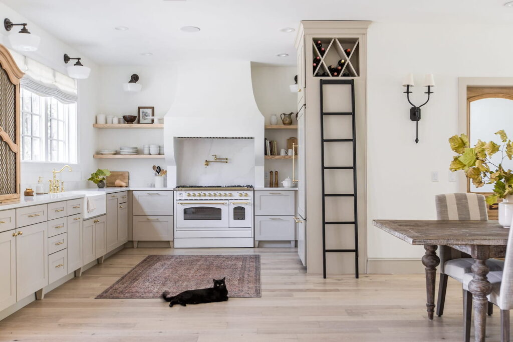A warm, French-inspired kitchen and living space with light gray cabinets, white appliances and light wood accents