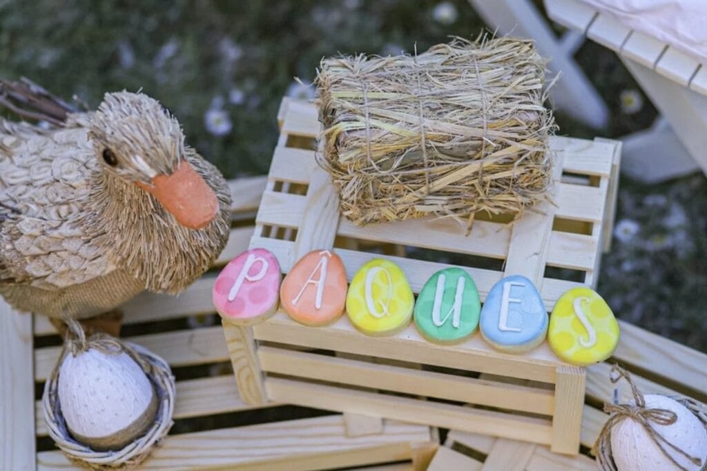 A wooden duck sits next to a small crate with a mini bale of hay and decorated cookies spelling out "Paques," which means Easter in French.