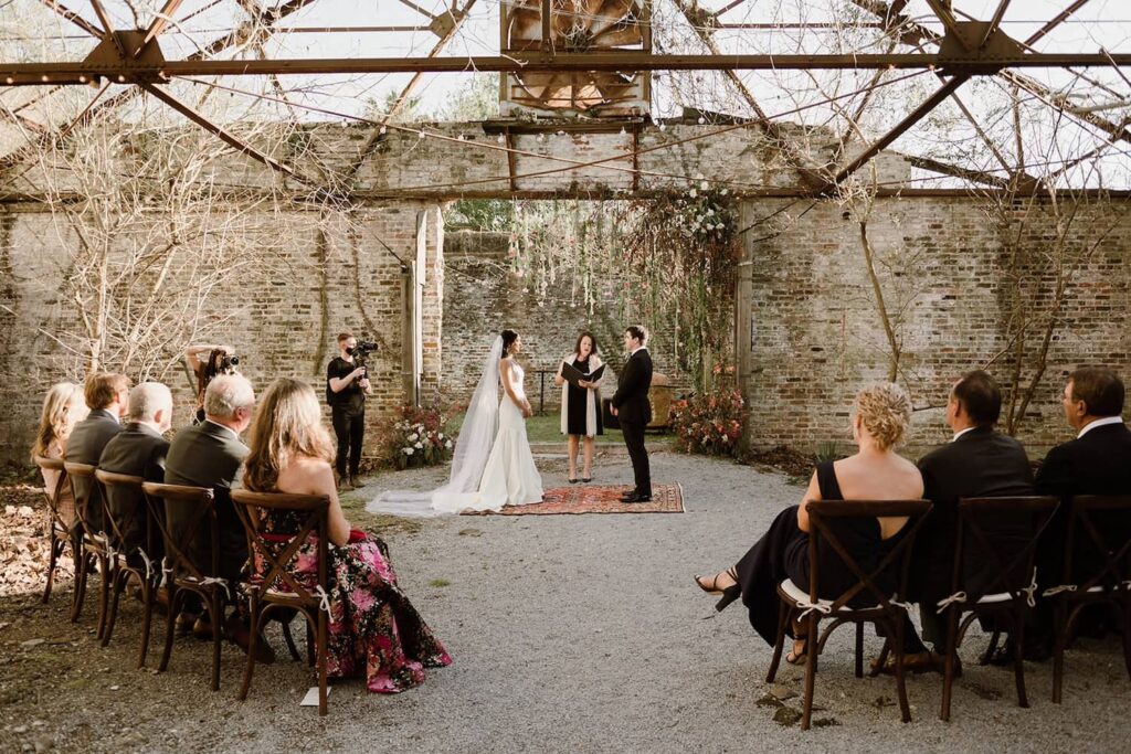 A bride and groom exchange vows in a rustic garden venue with brick walls and a gravel floor surrounded by a handful of friends and family