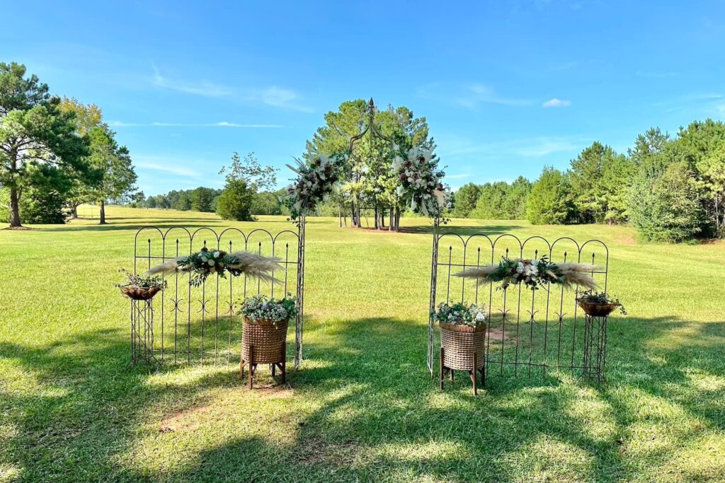 A small wrought iron gateway decorated with flowers leads to a large expanse of lawn on a farm