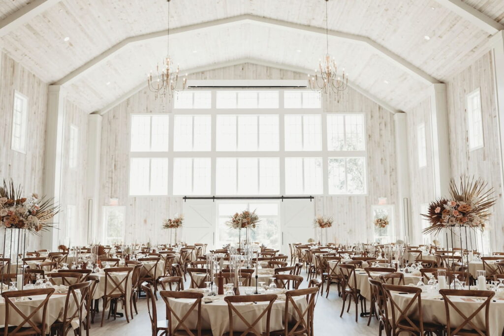 Whitewashed wooden barn interior with gigantic windows, white wooden barn doors and numerous round tables decorated with high and low boho-style wedding florals