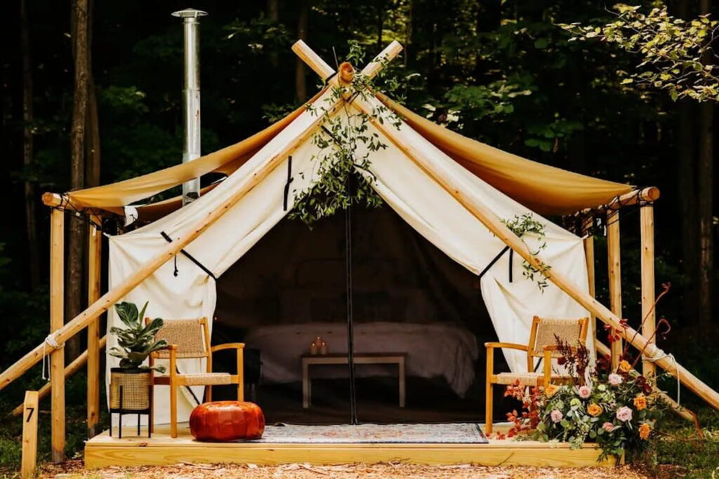 A glamping tent set up in the woods with candles lit inside and chairs on the porch surrounded by plants and floral arrangements