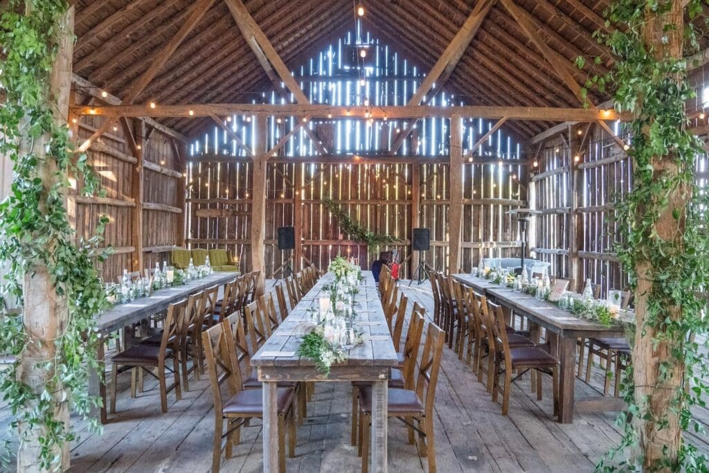 Three long wooden tables and chairs decorated with candles and greenery sit inside a quaint barn lit with string lights