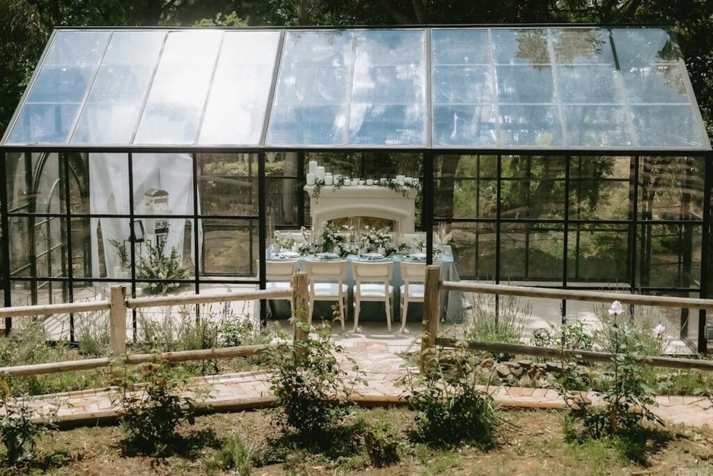 A small reception is set up in a greenhouse decorated with blue linens, greens and candles