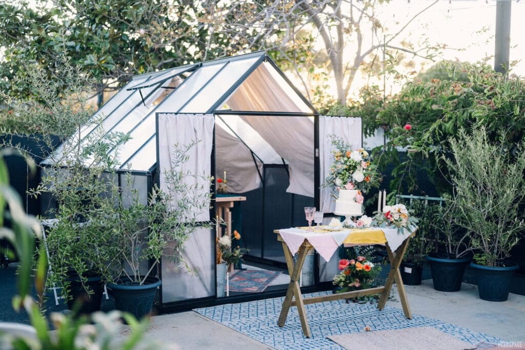 Wedding cake sits atop rustic table in front of a small greenhouse