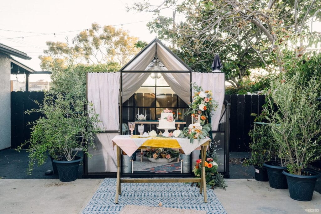 Wedding cake sits on wooden table in front of small, draped greenhouse surrounded by lush plants and flowers