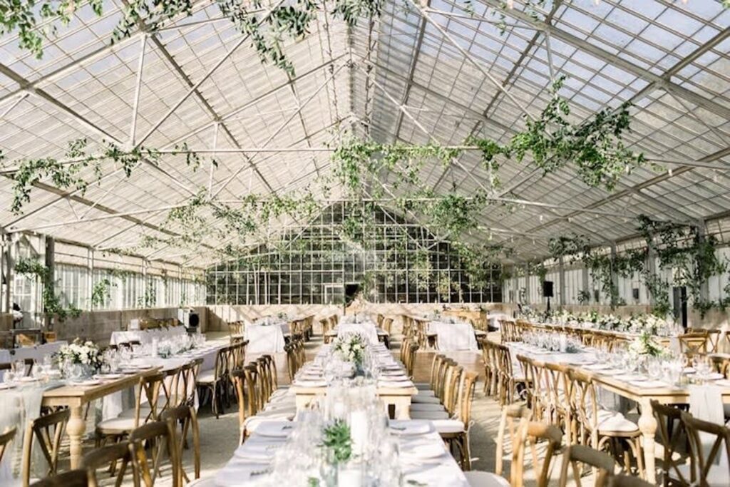 A bright greenhouse is set up for a wedding reception with weathered wooden tables and chairs decorated with neutral linens and white flowers