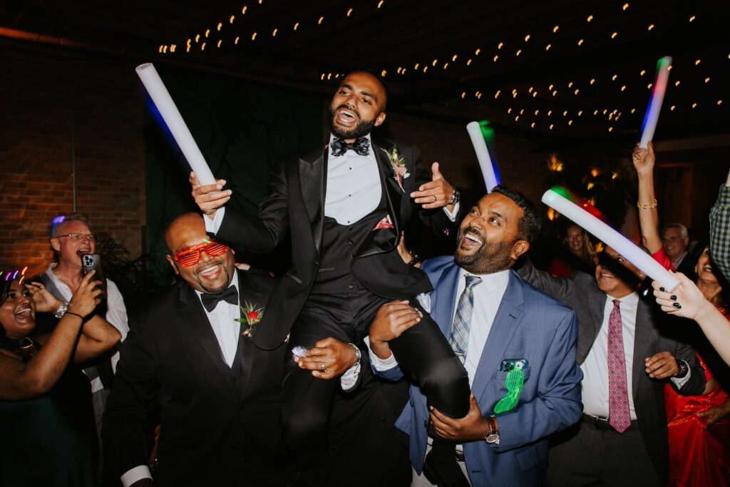 A groom is lifted up by his friends and family in celebration on the dance floor at his wedding.