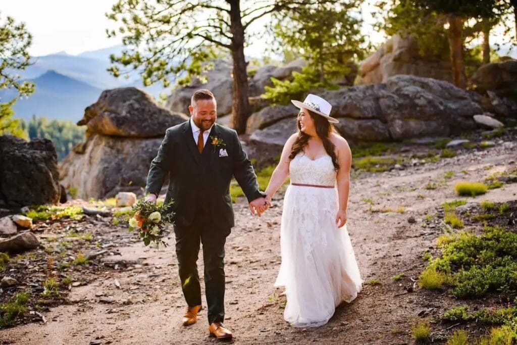 A bride and groom walk hand in hand down a dirt path with mountains in the distance as they smile at each other