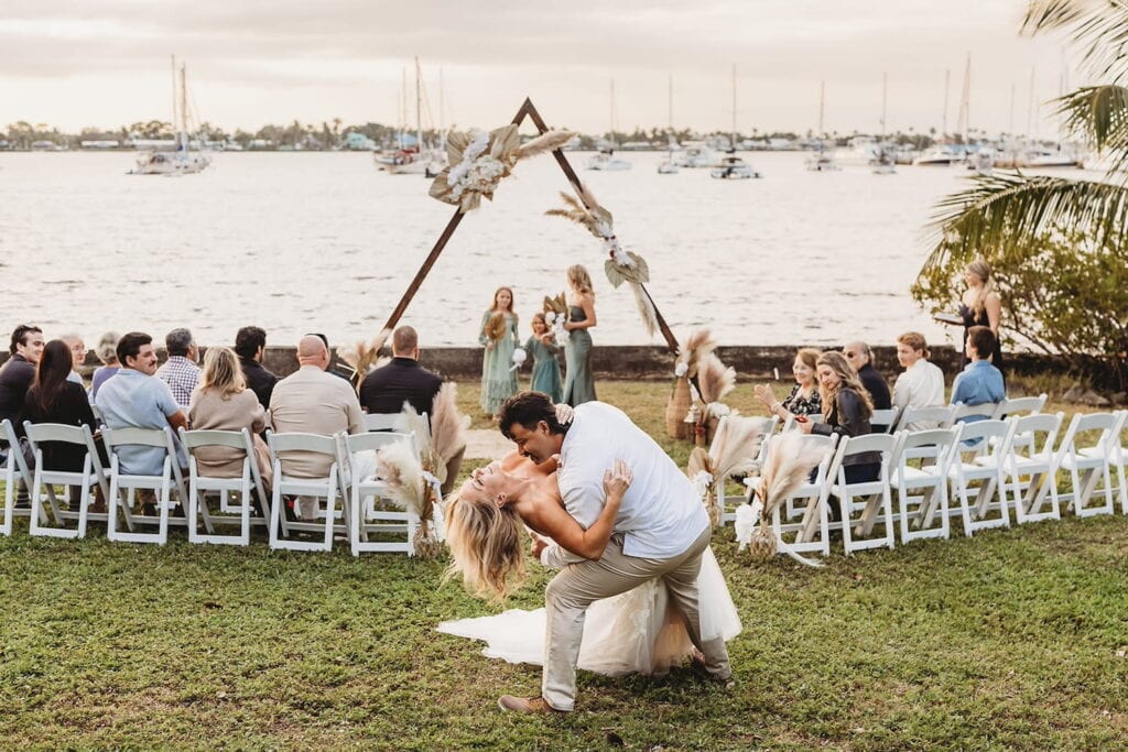 A groom dips his bride at the end an aisle at an outdoor wedding venue by the water