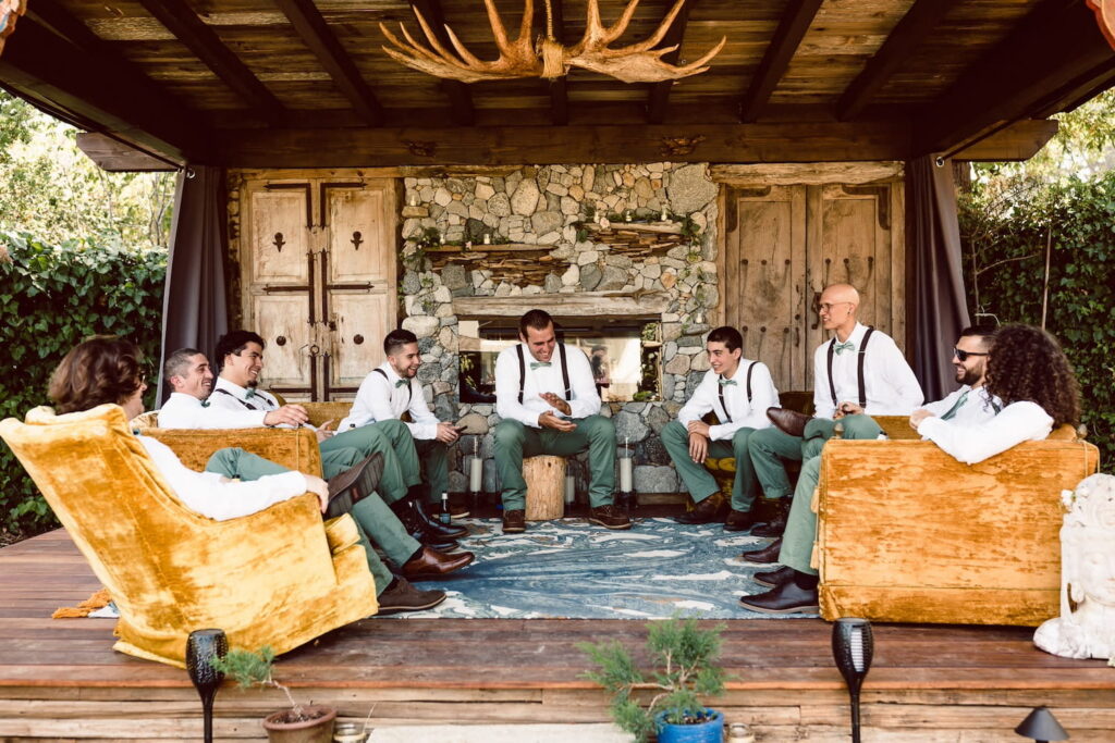 A groom and his wedding party share a moment in an outdoor living space with a stone fireplace and wood floors