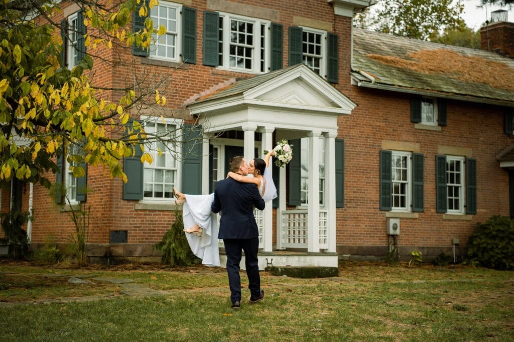A groom carries his bride towards a brick, colonial home while they share a kiss