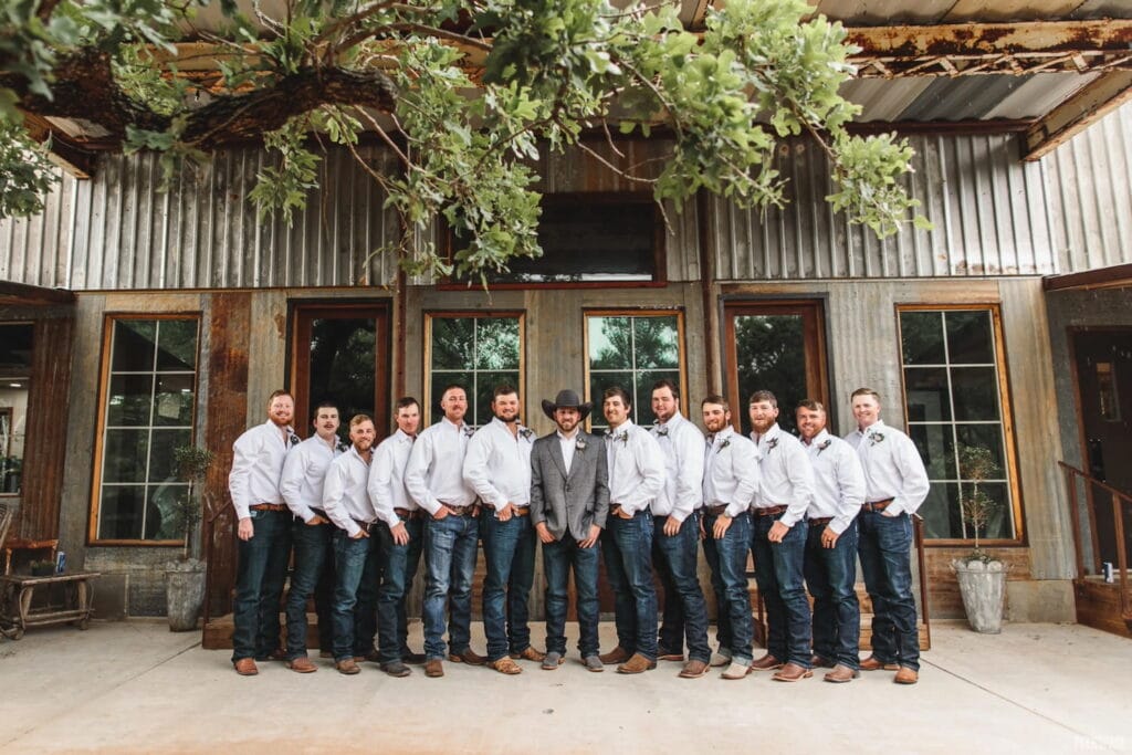 Twelve groomsmen in jeans, cowboy boots and button downs flank a groom in a cowboy hat