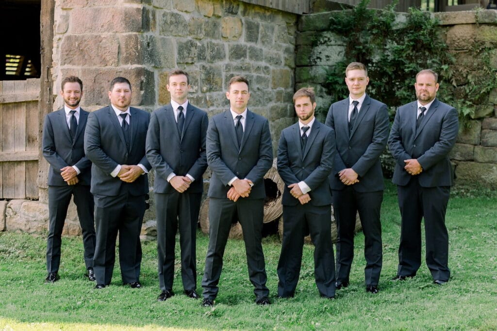 A row of groomsmen in black suits stand with a groom in front of a rustic wood and stone building