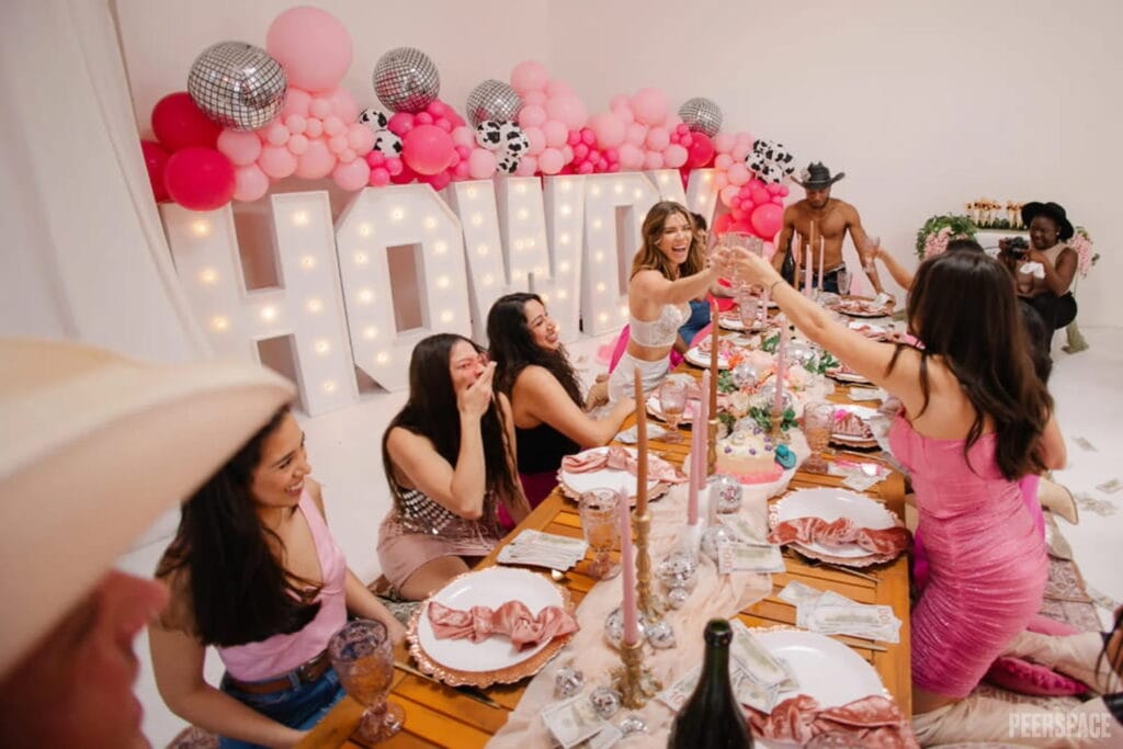 A group of women sit at a long table in front of a giant, light-up sign that says, "Howdy" and is covered in pink balloons