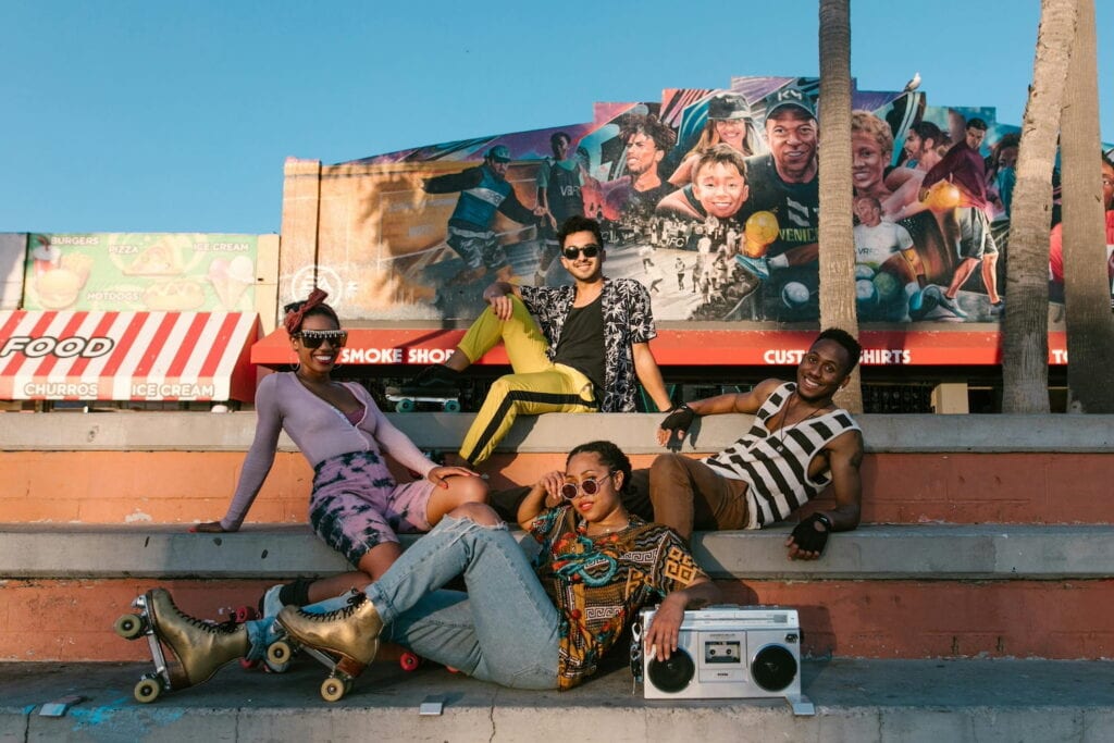 A group of people sit on cement steps in bright clothes wearing roller skates and listening to a boom box