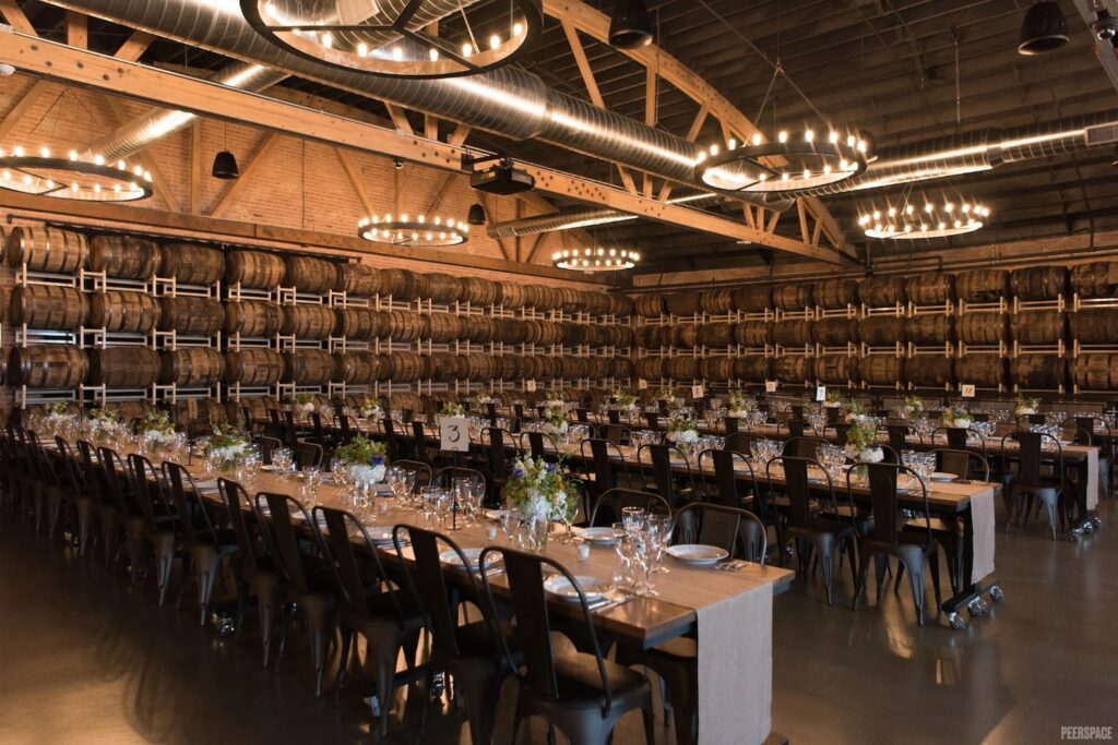 Four long guests tables are decorated simply with low-lying centerpieces and metal dining chairs in a room decorated with wine barrels and chandeliers