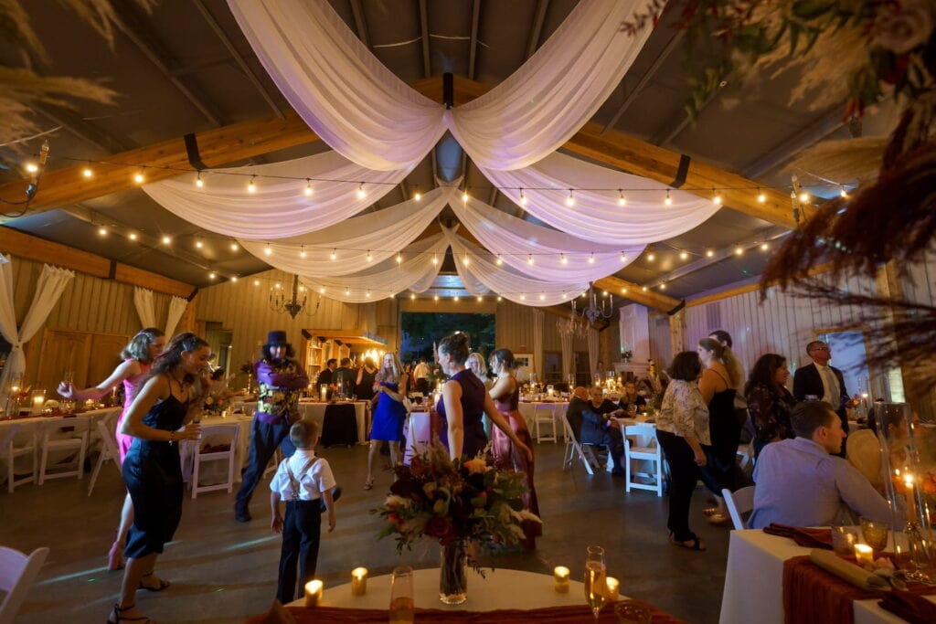Guests gather on a dance floor beneath string lights and chandeliers in a romantic, barn-like wedding venue