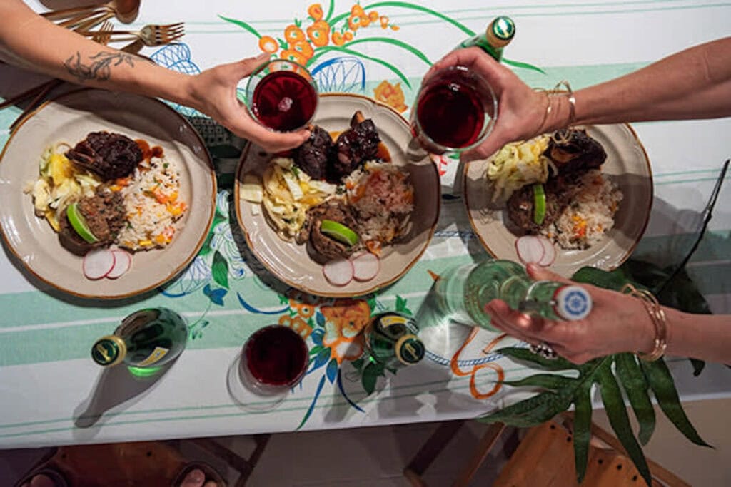 Overhead image of a table with plates of Hawaiian food and hands holding wine glasses reaching over in a toast