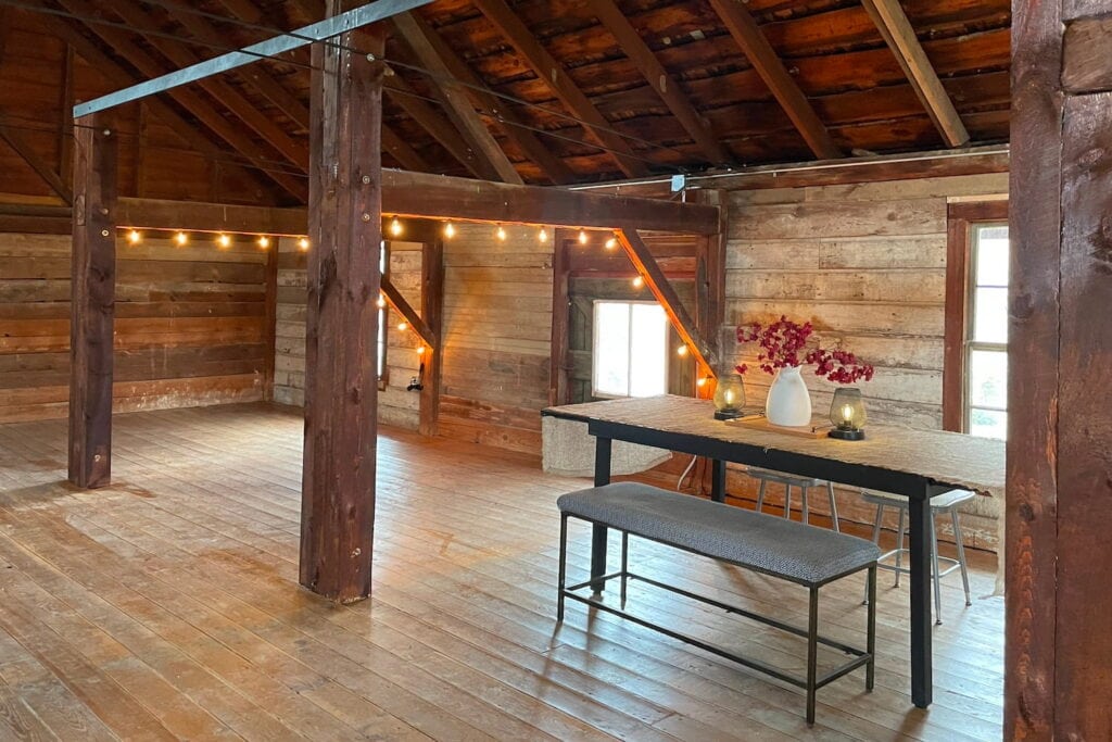 A rustic hayloft with string lights, large windows, and a small dining table lit with candles