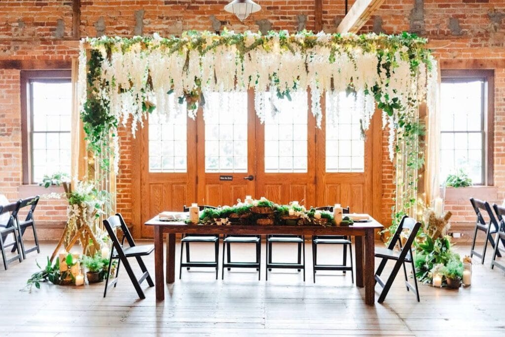 A wooden table with greenery and candles sits underneath a canopy of flowers by large wooden doors