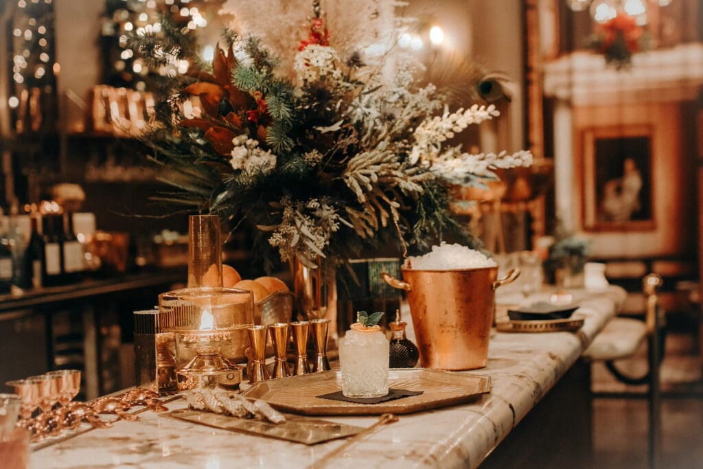 A lush centerpiece sits on a marble bar surrounded by ice buckets, shot glasses, and other cocktail accoutrements