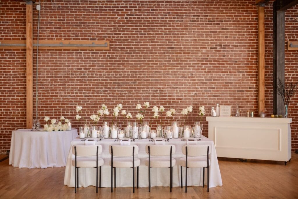 Exposed brick walls punctuate a venue space set up for a simple reception with all white tables lined with candles and white flowers