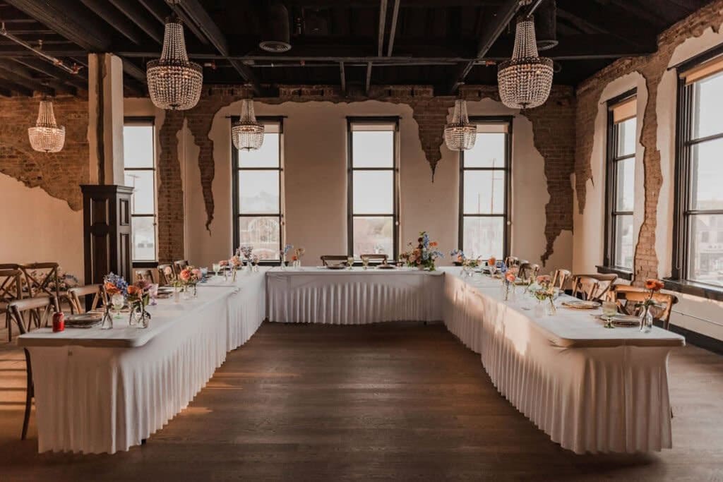 A U-shaped reception table is set in an historic venue with partially exposed brick walls, tall windows and chandeliers hanging from the ceiling