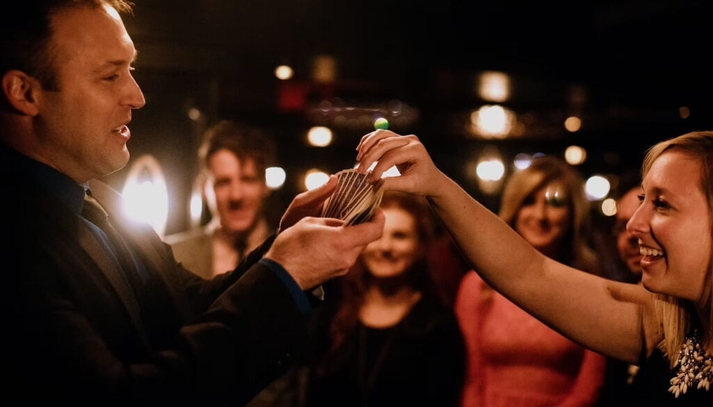 A woman selects a card from a card deck held by a magician in front of smiling onlookers
