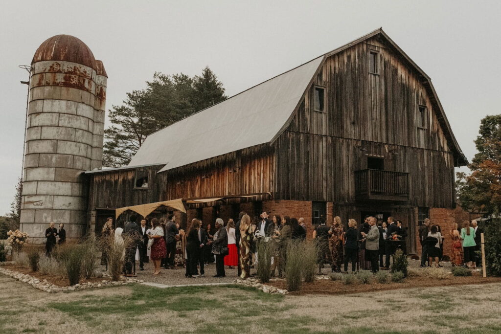 A crowd mills about outside a large wooden barn and attached silo in formal clothes