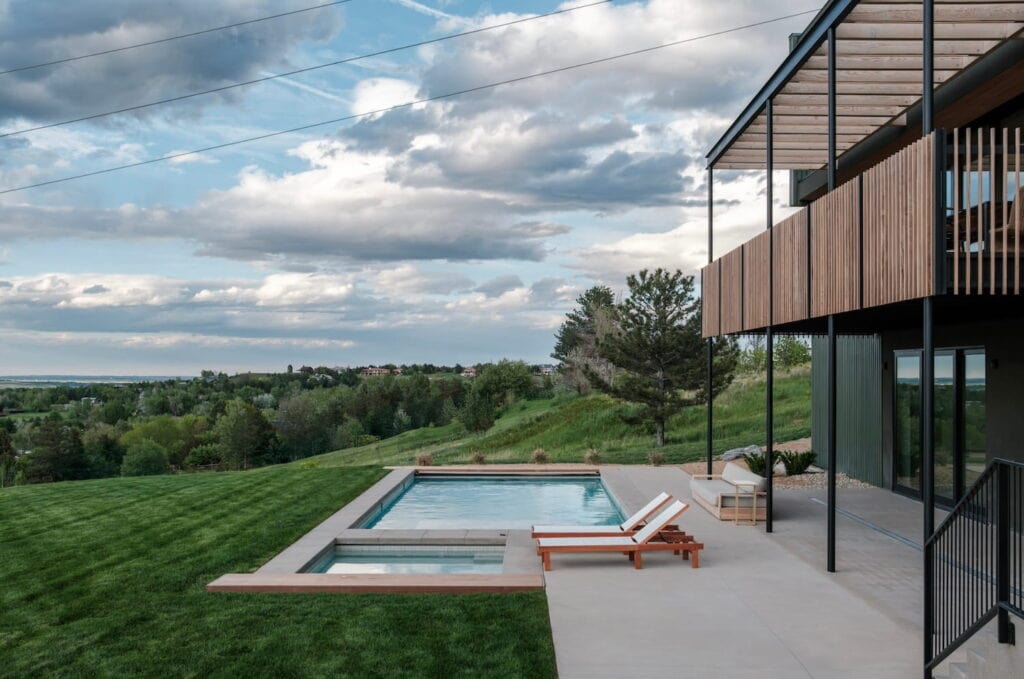A pool and hot tub sit next to a modern home overlooking rolling green hills in Colorado