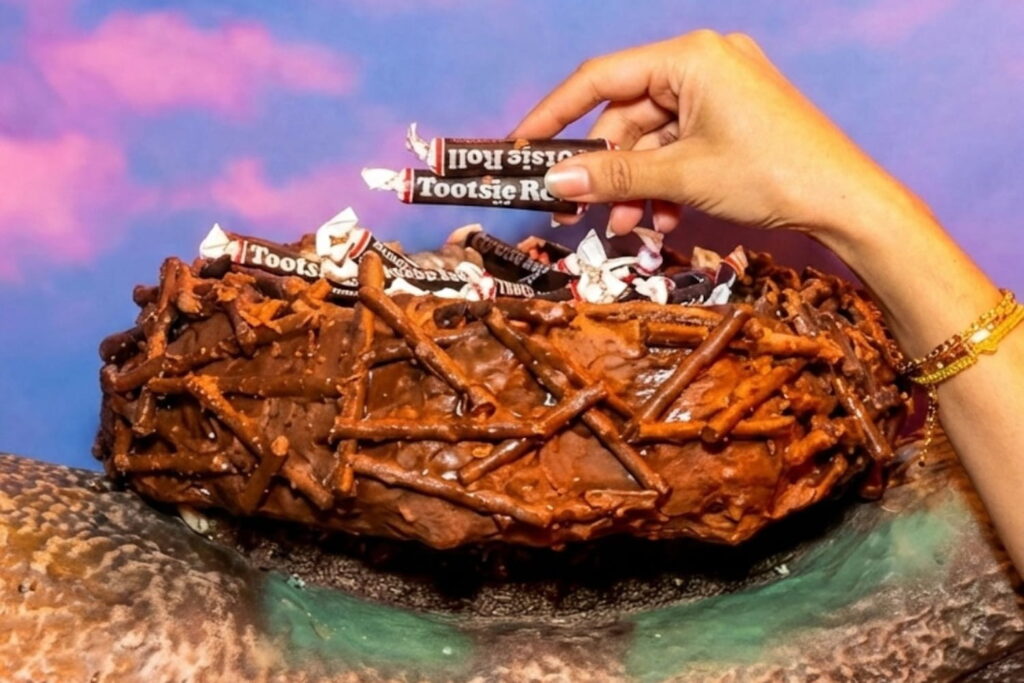 A hand holds two Tootsie Rolls above bow made of chocolate and pretzels