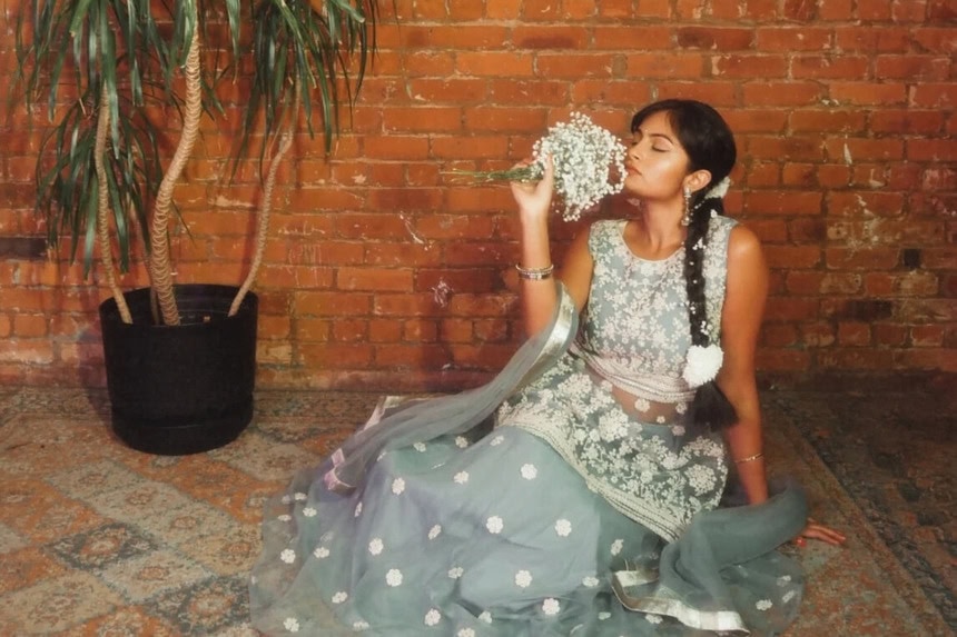 Woman in embellished South Asian attire posing with flowers against brick wall