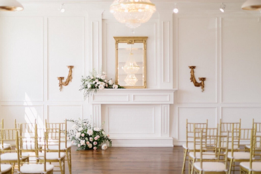 Gold chairs create an aisle facing a wooden mantle covered in flowers