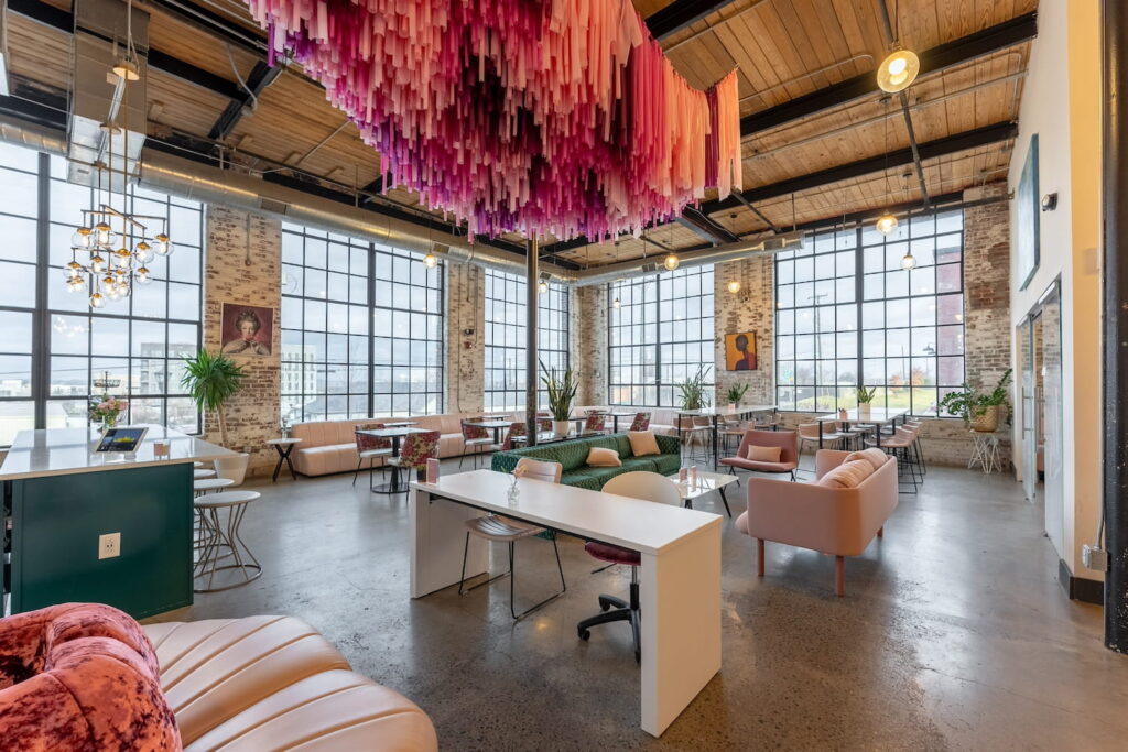 An industrial space with huge windows and brick walls is softened with velvet couches in pink and green, chandeliers overhead and a colorful art piece hanging from the ceiling
