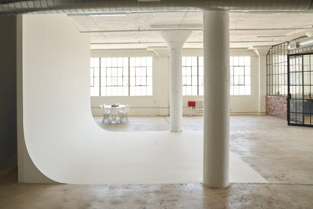 A small, round office table sits by large windows in an industrial venue with exposed pillars, concrete floors and brick walls