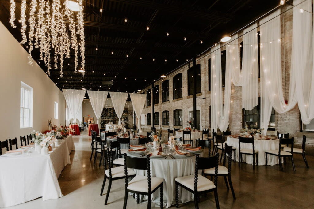 An bright industrial space is set up for a wedding reception with a long head table and multiple round guest tables